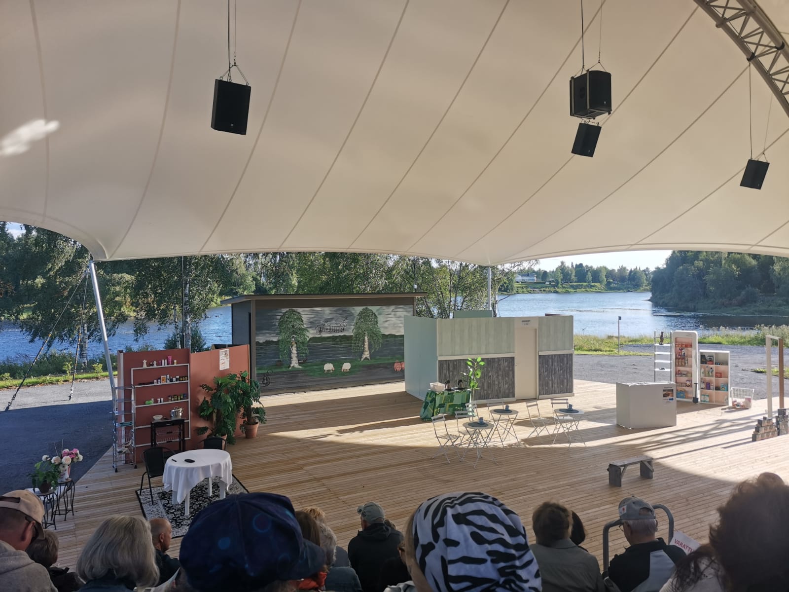 Väinölänrannan ulkoilmakatsomo, jonka päällä on valkoinen purjekatos. Katsomo on täynnä yleisöä ja taustalla näkyy kesäinen maisema.